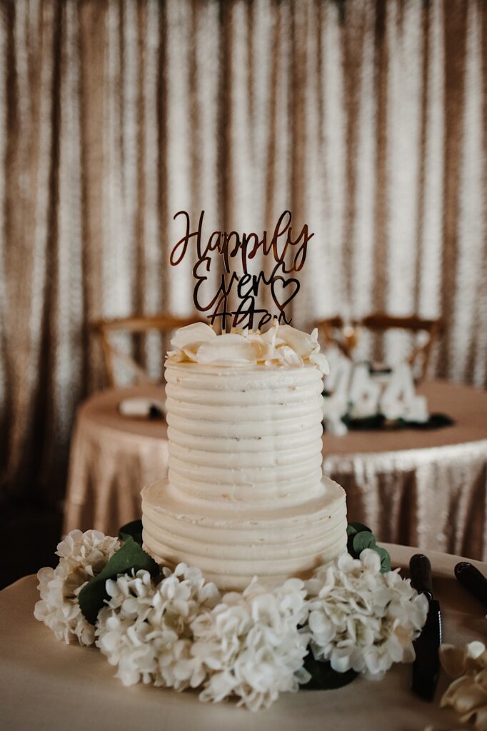 A wedding cake sits on a table surrounded by flowers with a sign on it that reads "Happily Ever After"