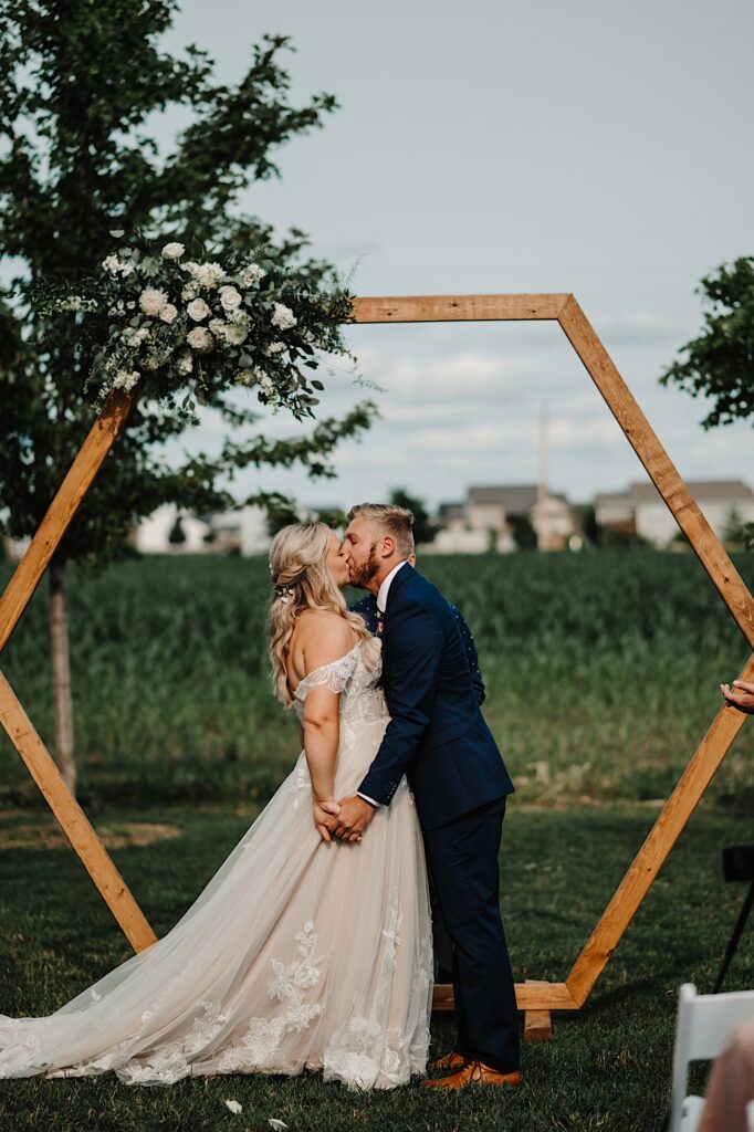 A bride and groom kiss underneath a wooden archway during their outdoor wedding ceremony