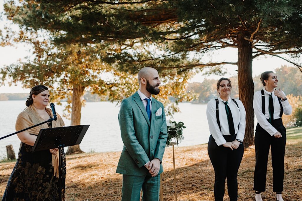 A groom stands next to members of his wedding party and his officiant in front of a lake during his fall intimate wedding reception near Bloomington Illinois