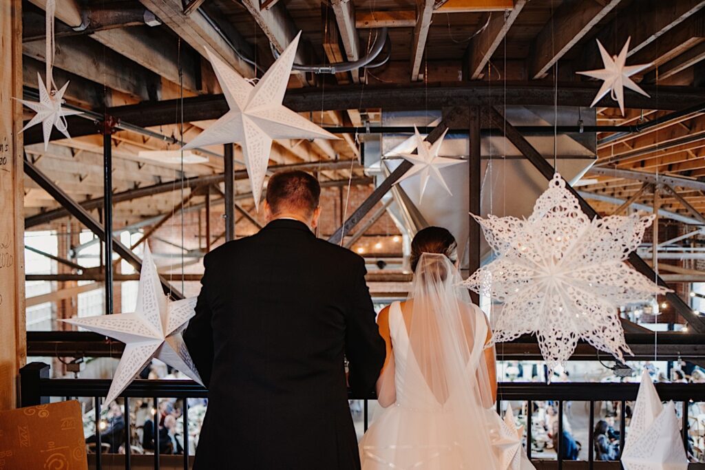 A bride and groom stand facing away from the camera looking out from an indoor balcony over the reception space below them at Stardust in Davenport, in front of them hanging from the ceiling are large paper stars