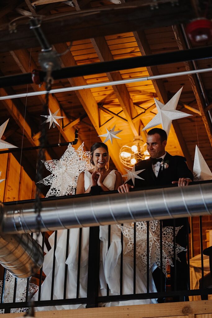 A bride and groom stand next to one another on an indoor balcony and look over a railing, hanging from the ceiling around them are large paper stars