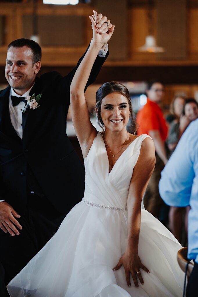 A bride and groom smile while walking next to one another and lifting their arms in the airs as they enter their wedding reception