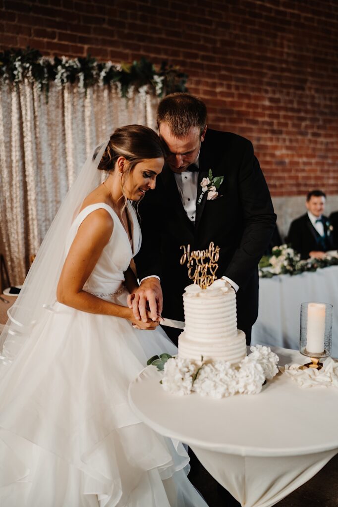 A bride and groom stand next to one another and cut their wedding cake together during their wedding reception