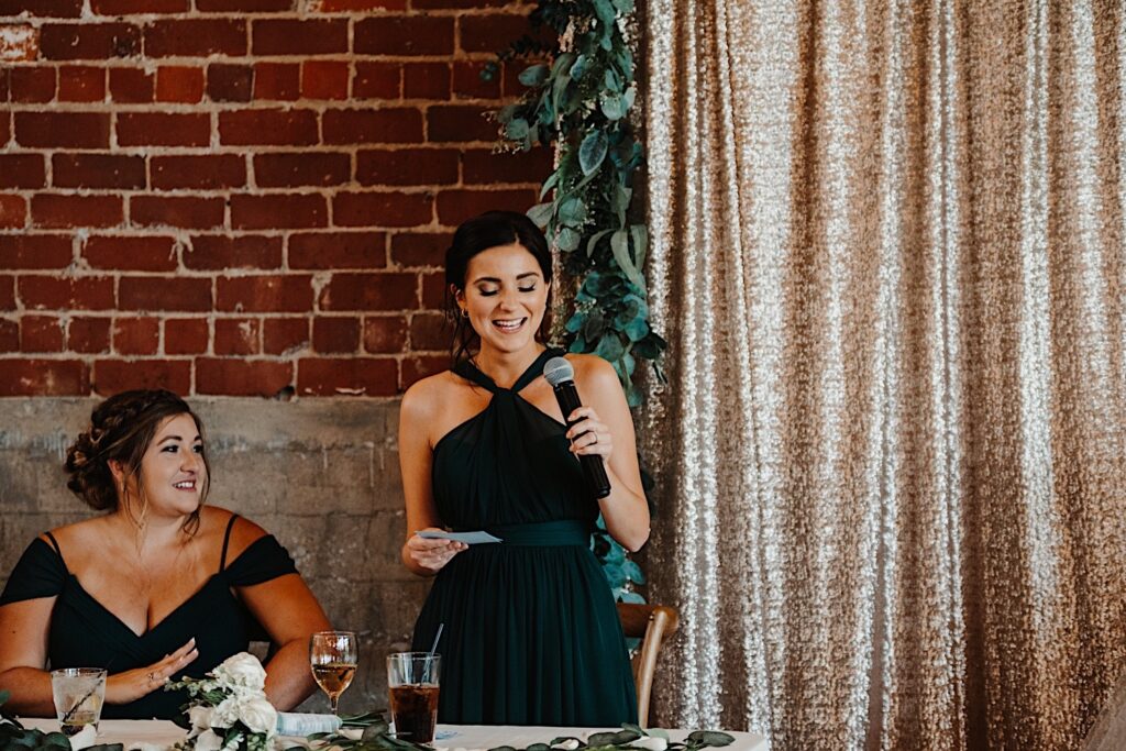 A bridesmaid smiles while standing and giving a speech during an indoor wedding reception at Stardust in Davenport as another bridesmaid to the left of her smiles at her