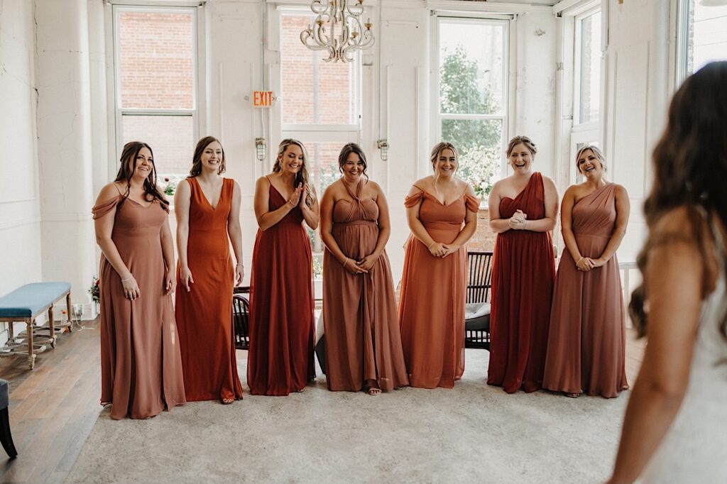Seven bridesmaids all stand and smile in a well lit room as a bride in her wedding dress stands in the foreground of the photo for the first look, photographed by a Central Illinois wedding photographer