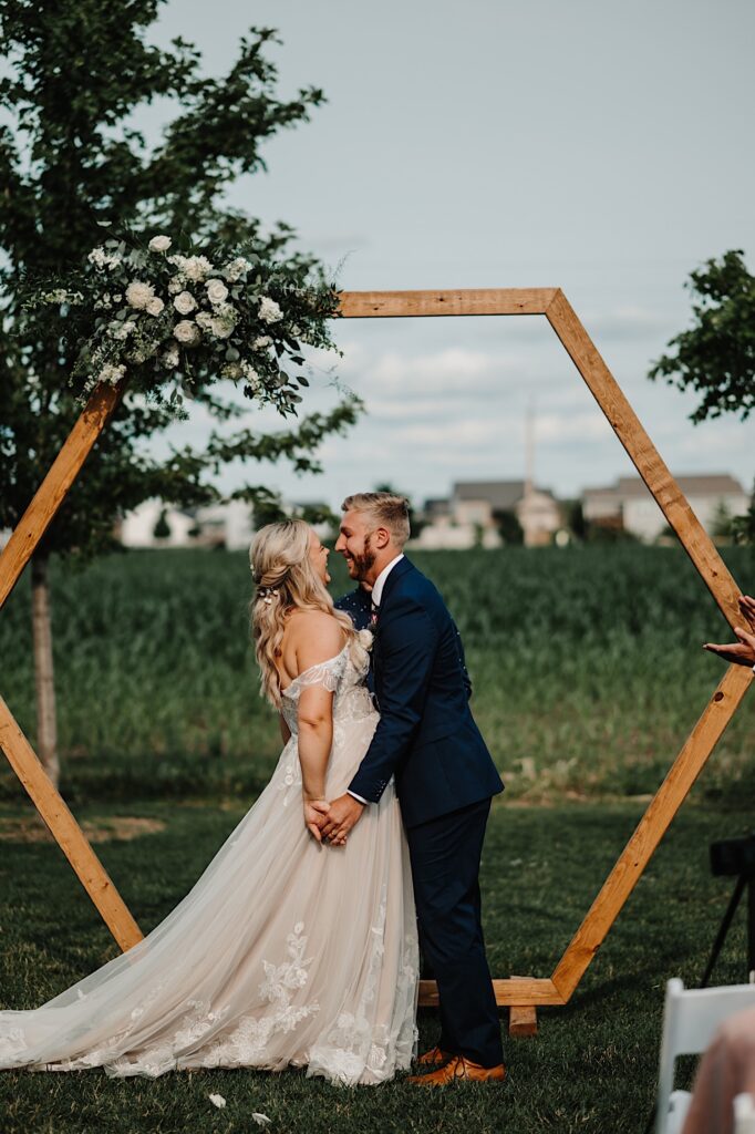 A bride and groom smile after kissing underneath a wooden arch during their outdoor wedding ceremony