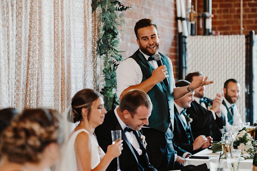 A bride and groom are laughing while sitting as a groomsman to the right of them stands and gives a speech during their indoor wedding reception at Stardust in Davenport