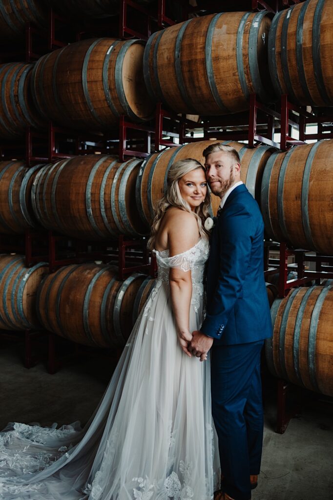 A bride and groom hold hands and face one another while smiling at the camera in front of a wall of wooden bourbon barrels