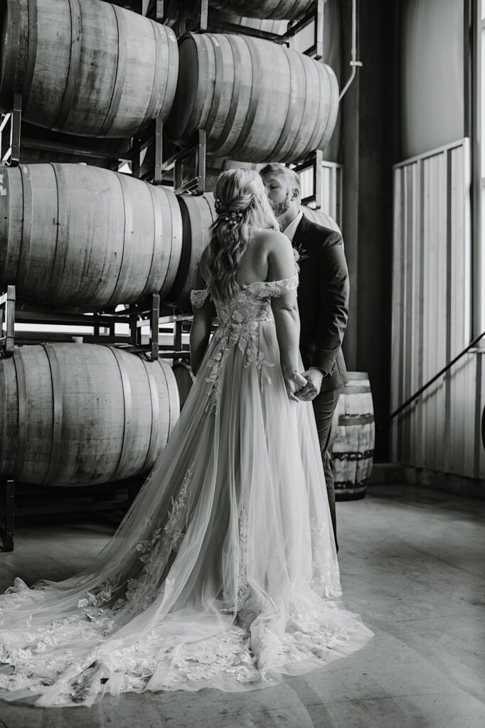 Black and white photo of a bride and groom holding hands while kissing one another in front of a wall of bourbon barrels