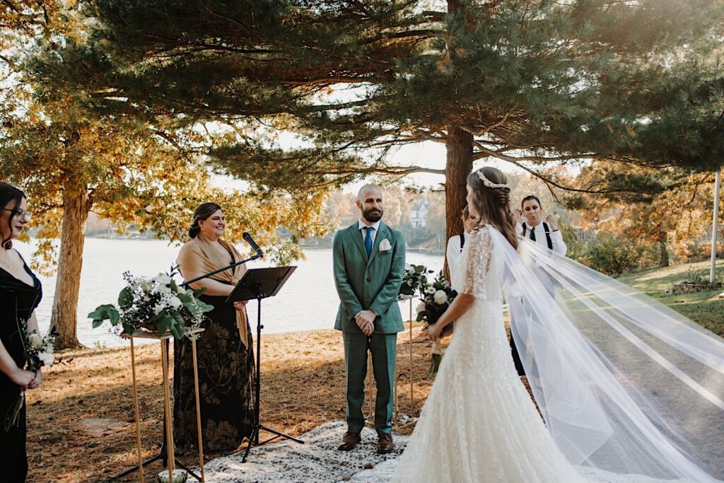 A groom smiles at the bride as she walks down the aisle as their officiant and wedding parties watch and smile as well, the intimate wedding is taking place in front of a lake near Bloomington Illinois in the fall