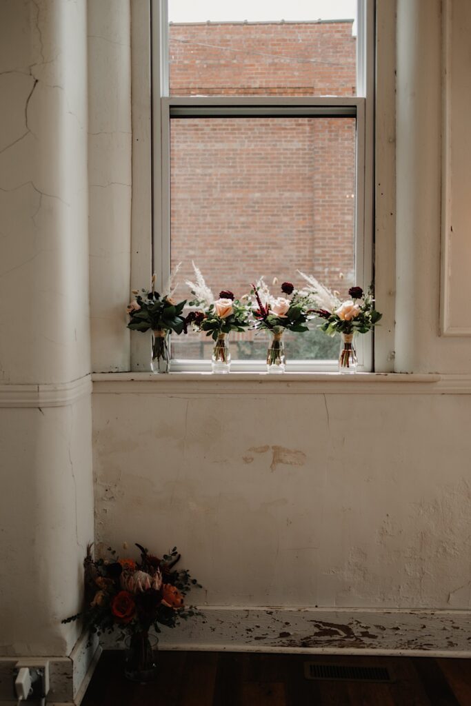 Four vases with flowers in them sit on a windowsill while a larger bouquet in a vase sits on the floor below