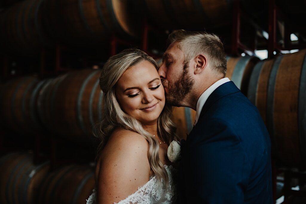 A bride smiles at the camera and closes her eyes as the groom kisses her cheek in front of a wall of bourbon barrels inside their wedding venue, Destihl Brewery in Normal, Illinois