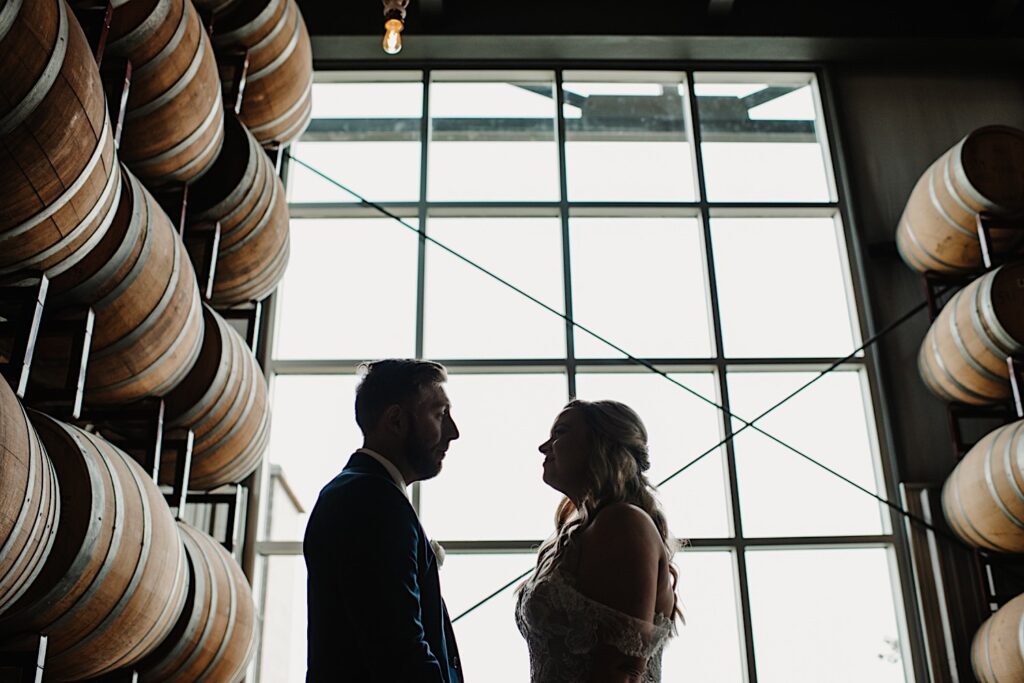 A bride and groom look at one another in front of a large window and with bourbon barrels on either side of them at their wedding venue, Destihl Brewery in Normal, Illinois