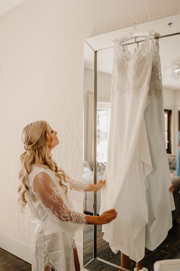 A woman touches her wedding dress that is hanging on a large mirror in front of her