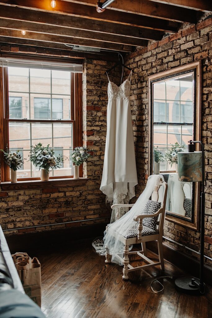 In a brick getting ready space a wedding dress hangs from the ceiling and a veil is draped over a chair
