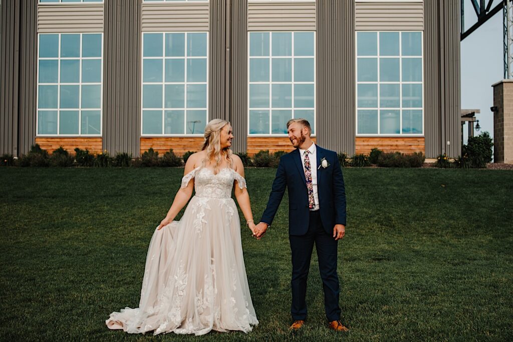 A bride and groom hold hands and smile at one another while facing the camera outside of their wedding venue, Destihl Brewery in Normal, Illinois