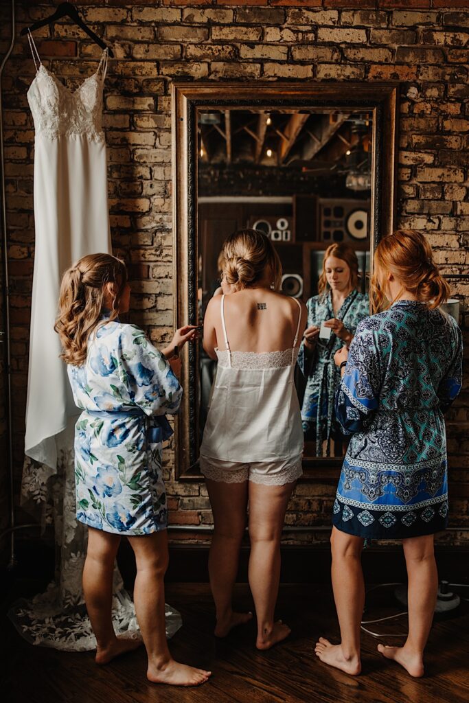 In a brick room a bride stands in the mirror putting earrings on while a bridesmaid on either side helps her