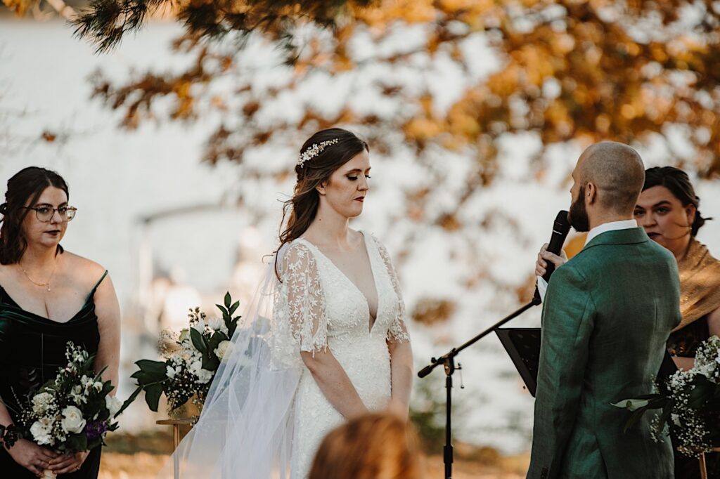 A bride looks down while the groom stands in front of her holding a microphone and says his vows as a bridesmaid and their officiant watch