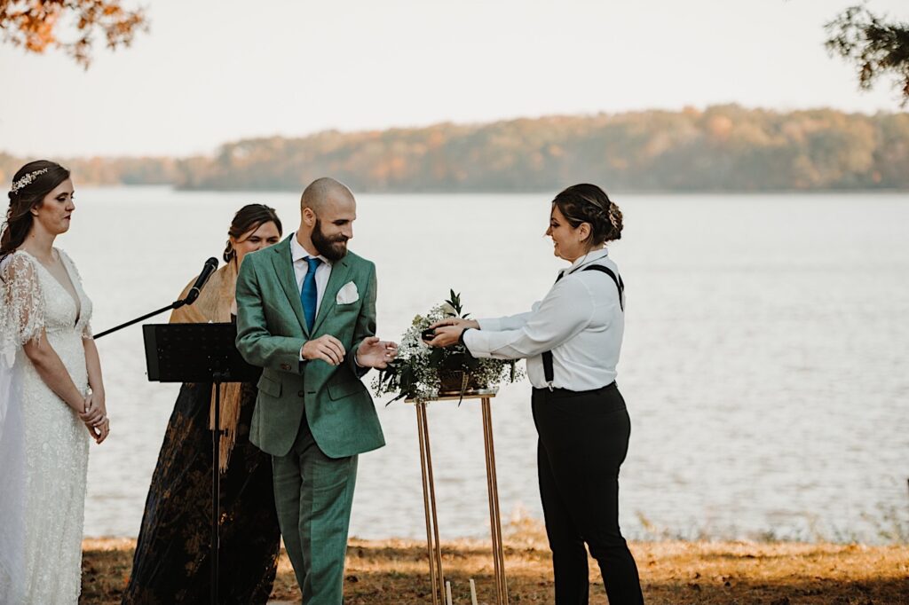 A groom smiles as he turns around to grab a wedding ring form one of his wedding party members during an intimate fall wedding ceremony in front of a lake near Bloomington Illinois