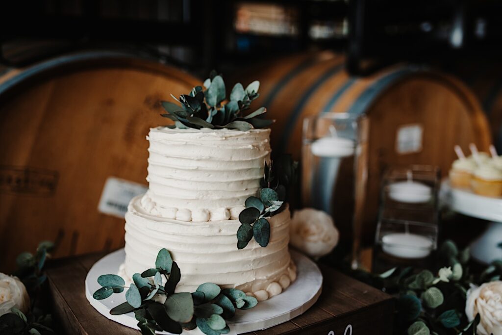 A white wedding cake with flowers on it sits on a table in front of bourbon barrels