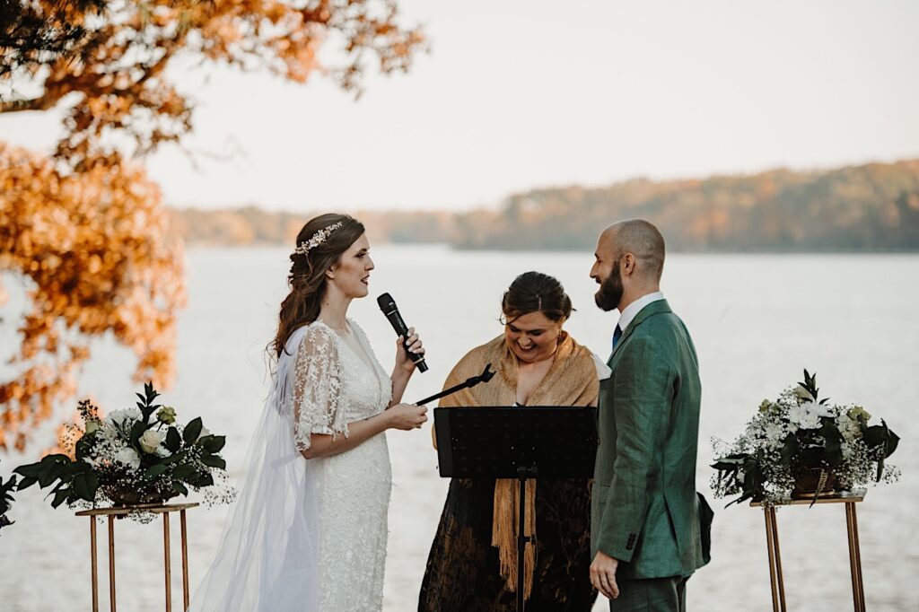 A bride holds a microphone and says her vows to the groom as he and their officiant laugh while standing in front of a lake in the fall