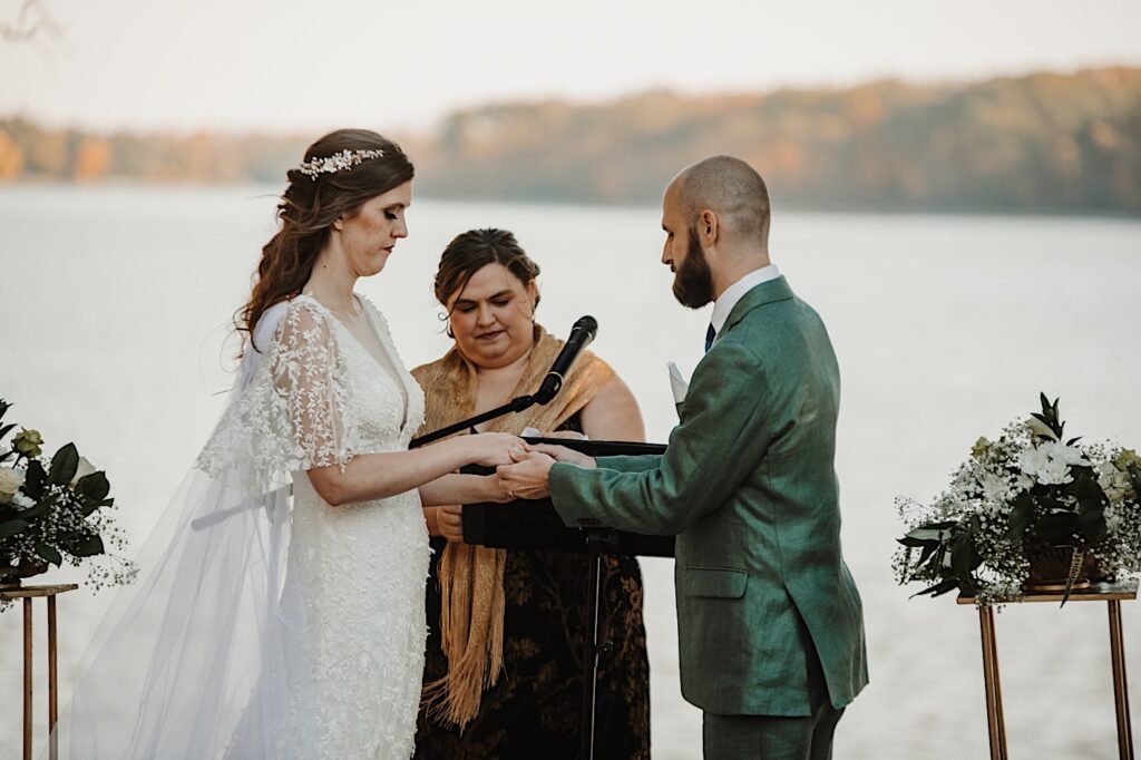 A groom puts a wedding ring on the bride's finger as their officiant watches during their wedding ceremony in front of a lake