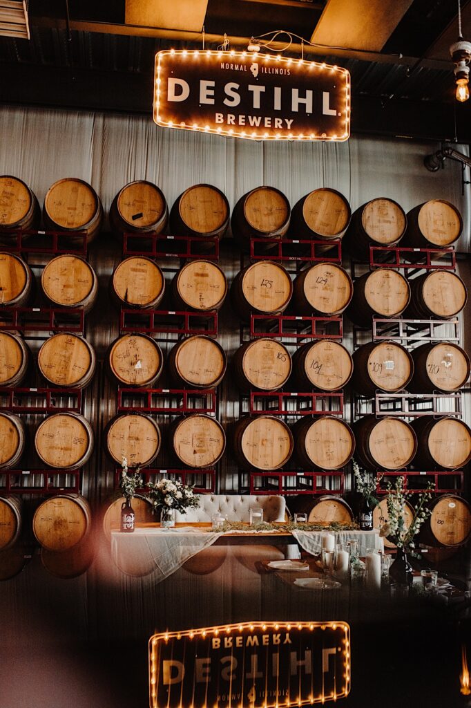 A wall of bourbon barrels underneath a sign for Destihl Brewery with the head table of a wedding set up in front of the wall