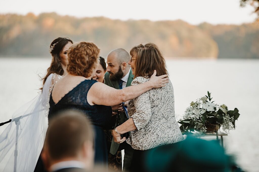 A groom smiles as two guests of the wedding come up to hug him and the bride during their wedding ceremony in front of a lake