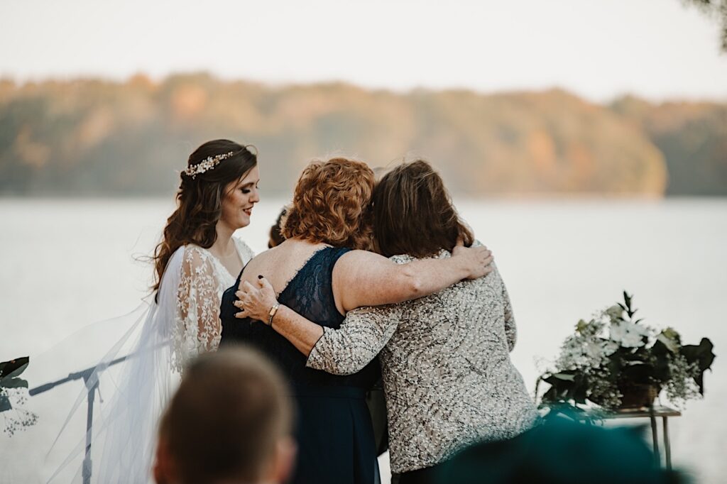 2 guests of a wedding hug one another next to the bride during a wedding reception in front of a lake