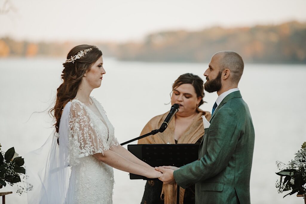 A bride and groom hold hands while looking at one another standing in front of a lake near Bloomington Illinois during their intimate wedding reception in the fall as their officiant speaks