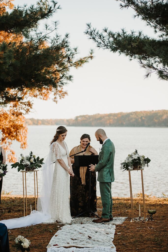 An officiant laughs while giving a speech during a wedding ceremony in front of a lake as both the bride and groom laugh in front of her