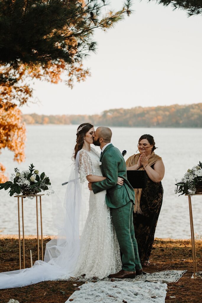 A bride and groom kiss while in front of a lake during their wedding ceremony as their officiant stands behind them and claps