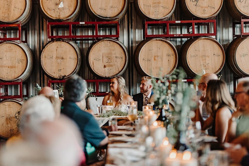 A bride and groom sit at the head table in front of a wall of bourbon barrels watching a speech during their wedding reception at Destihl Brewery's indoor venue space
