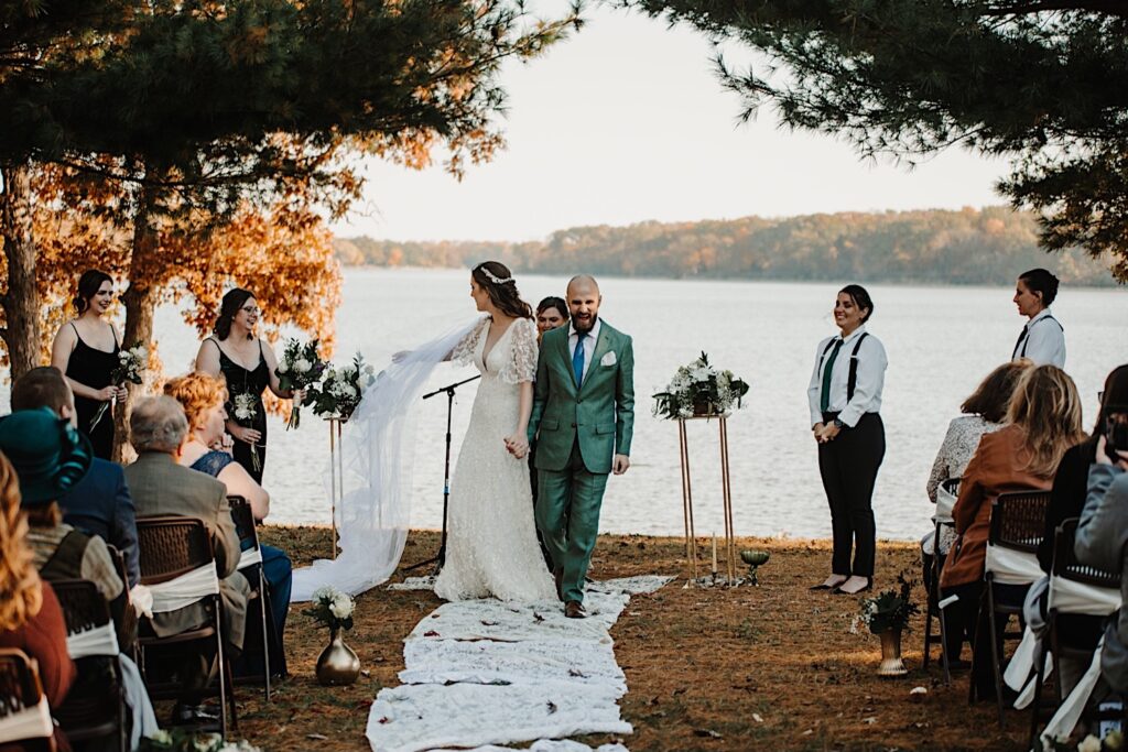 After their intimate fall wedding ceremony next to a lake near Bloomington Illinois a groom exclaims while holding the brides hand and walking down the aisle as the bride reaches back to her bridesmaid for her bouquet