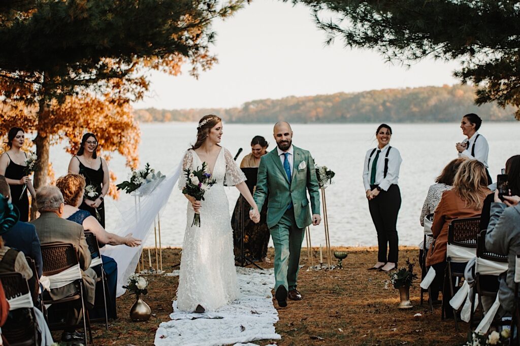 A bride and groom walk hand in hand down the aisle after their intimate fall wedding ceremony next to a lake near Bloomington Illinois as their wedding parties and guests on either side of them clap for them
