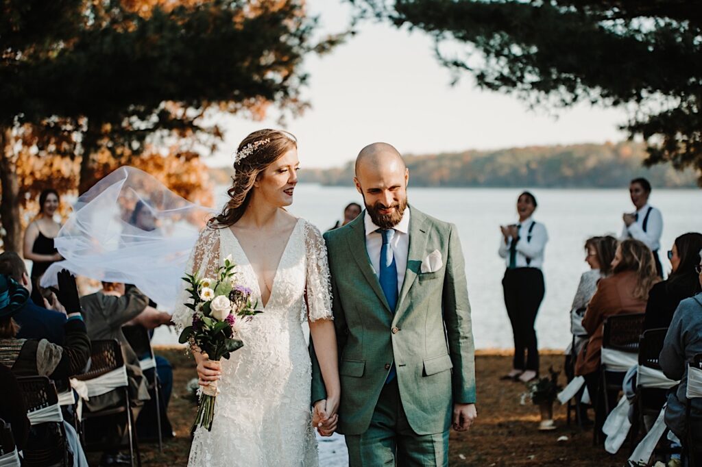 A bride and groom walk hand in hand down the aisle after their intimate fall wedding ceremony next to a lake near Bloomington Illinois as their wedding parties and guests on either side of them clap for them
