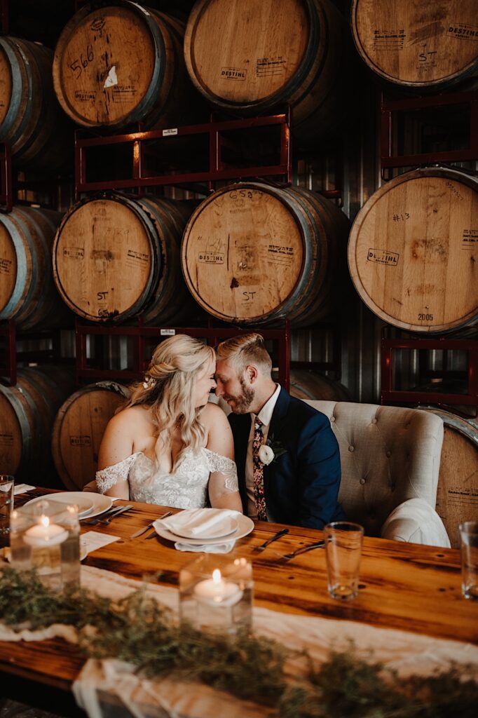 A bride and groom at the head table of their reception touch foreheads together and smile in front of a wall of bourbon barrels