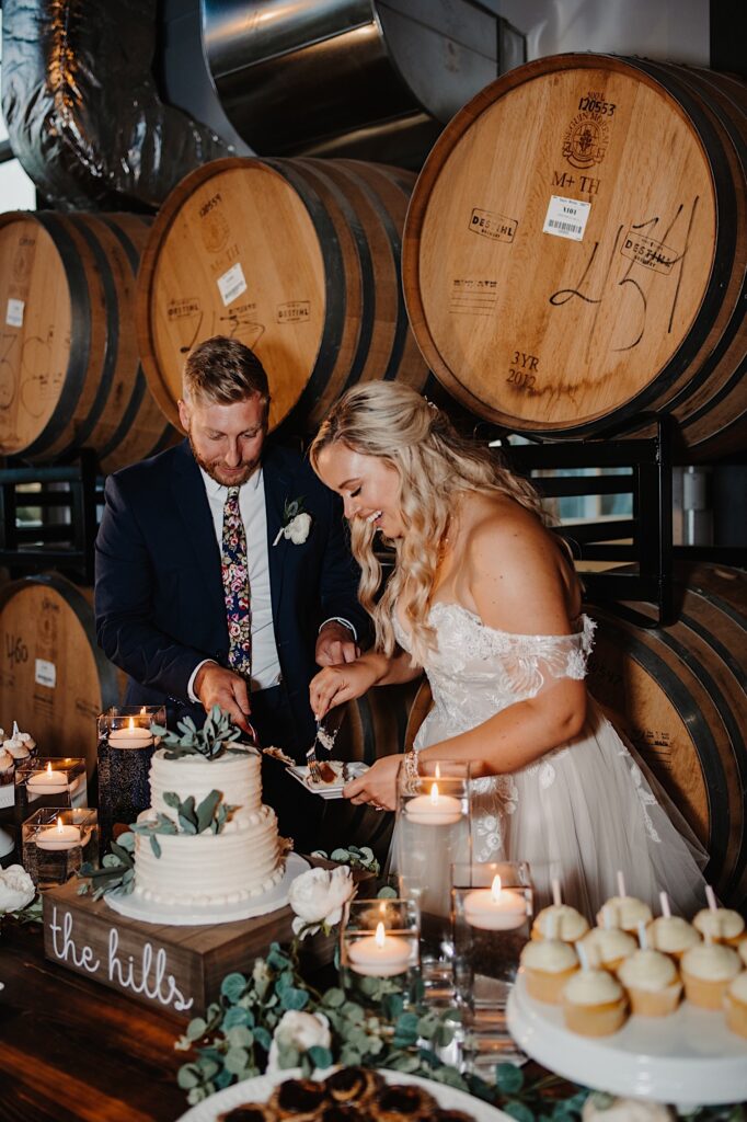 A bride and groom smile as they stand in front of a wall of bourbon barrels and cut their wedding cake together
