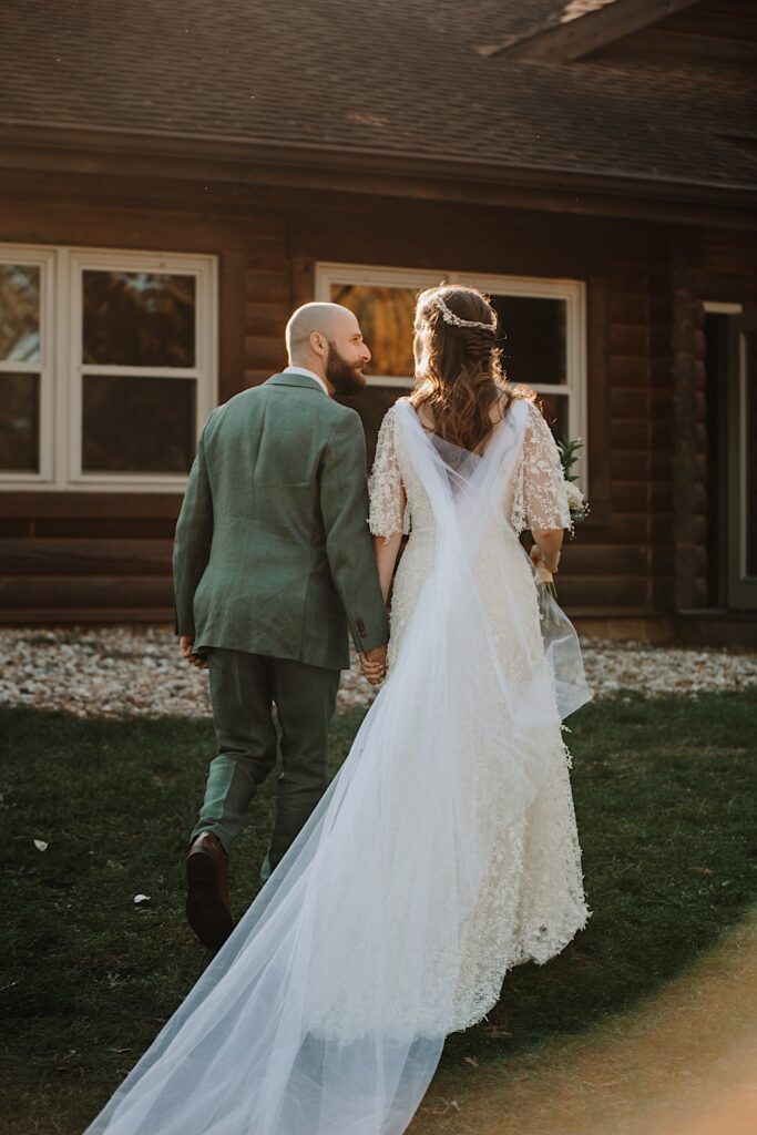 A bride and groom walk hand in hand towards a log cabin while looking at one another after their wedding ceremony