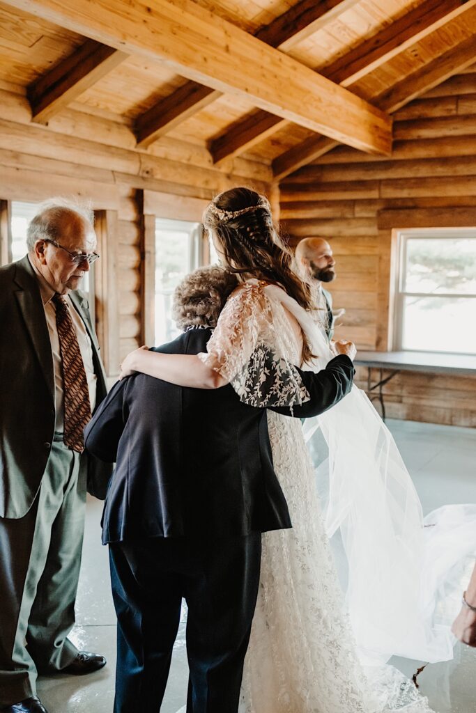 A bride hugs her grandmother while her grandfather stands next to them while all of them are inside a log cabin