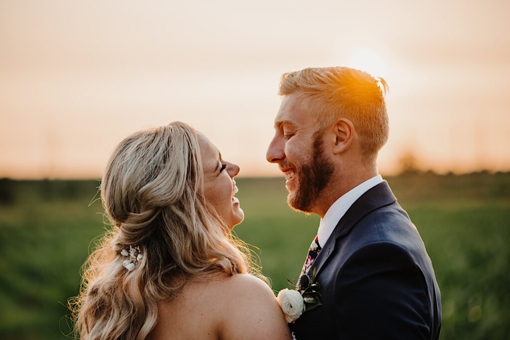 A bride and groom smile at one another and laugh as the sun sets behind them