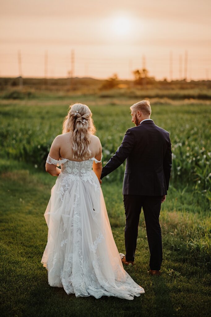 A bride and groom hold hands and walk away from the camera towards the sunset while in a cornfield