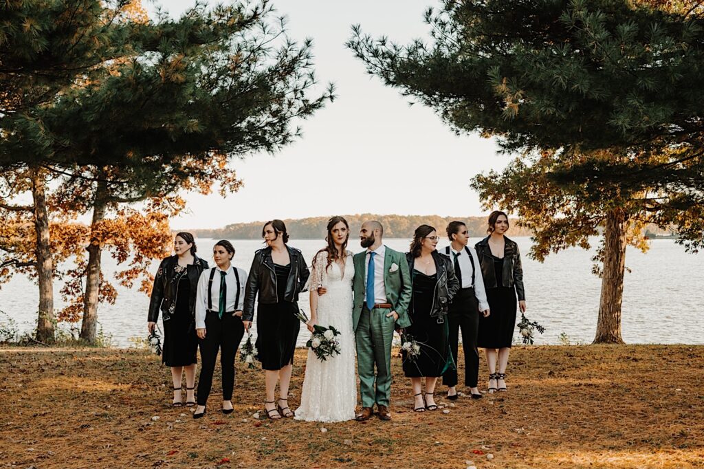 A bride and groom stand next to one another while the 6 members of their wedding parties stand on either side of them posing for the photo, behind them is a lake