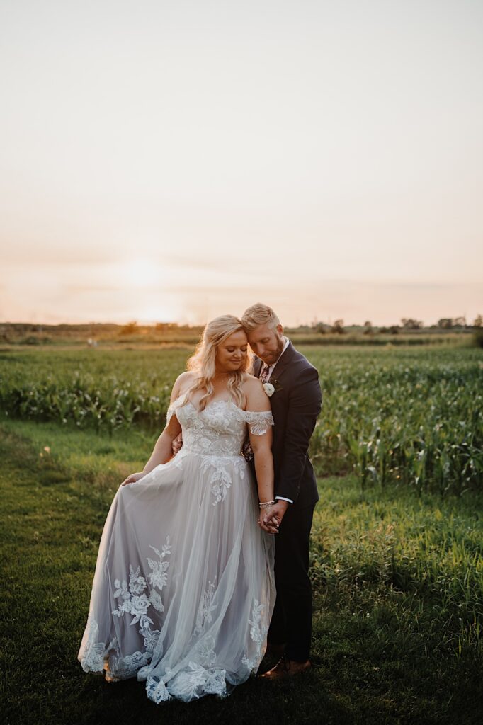 A groom stands behind the bride and holds her hand as they both look down while standing in a cornfield during sunset