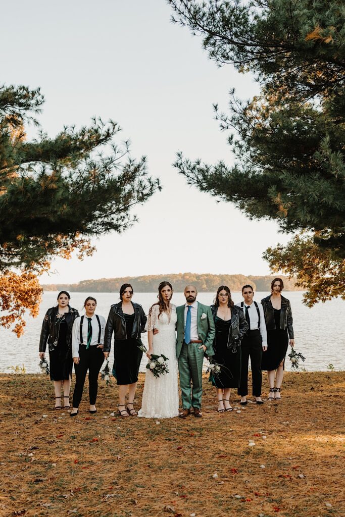 A bride and groom stand next to one another while the 6 members of their wedding parties stand on either side of them posing for the photo, behind them is a lake