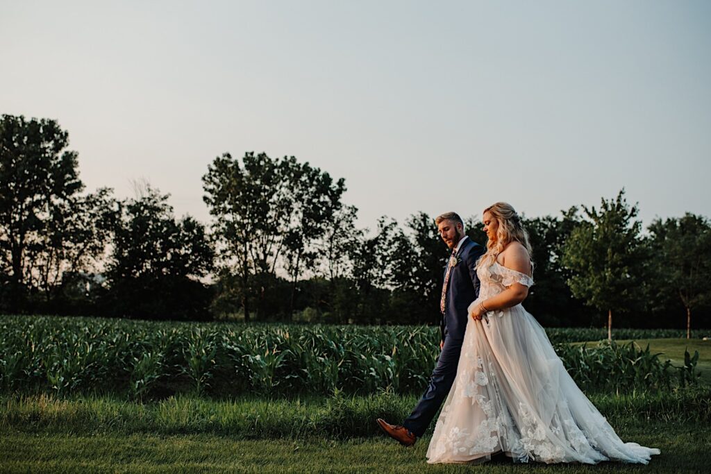 A bride and groom walk side by side next to a corn field outside of their wedding venue, Destihl Brewery in Normal, Illinois