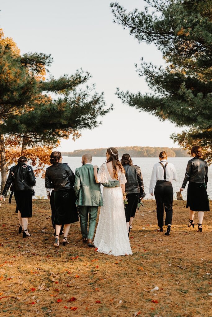 A bride and groom walk away from the camera with members of their wedding party on either side of them towards a lake