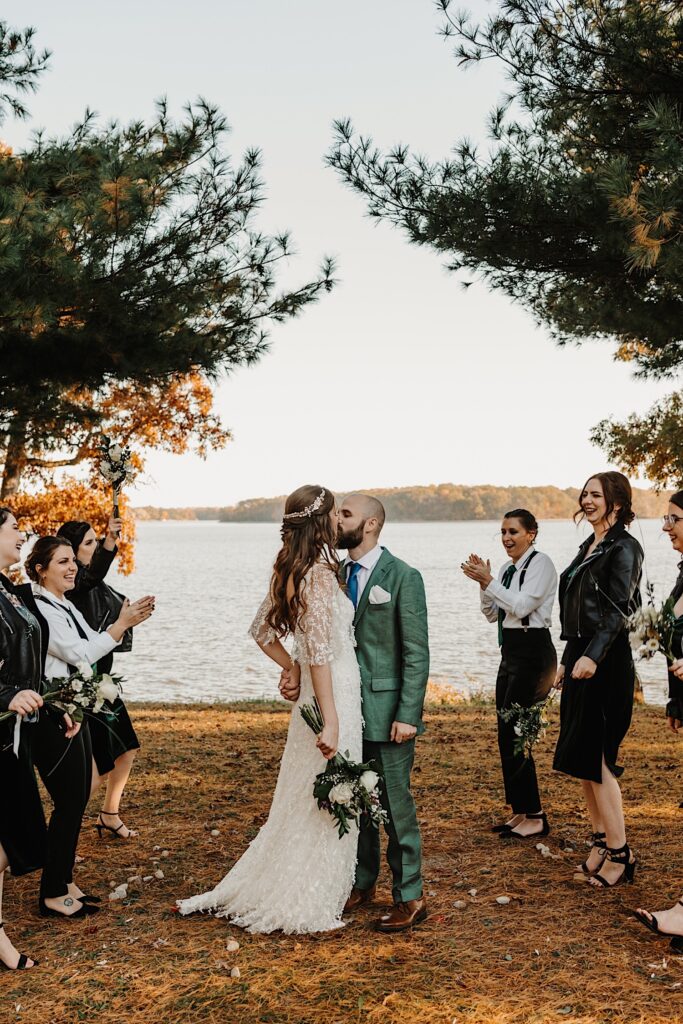 A bride and groom kiss one another in front of a lake while their wedding party members on either side of them cheer them on