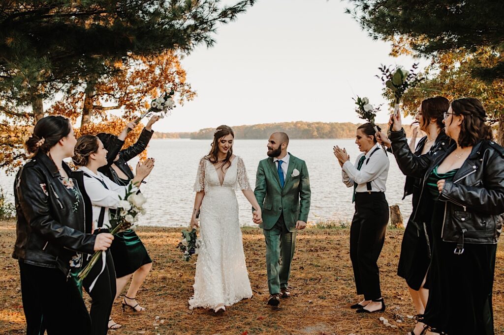 A bride and groom walk hand in hand towards the camera and smile while members of their wedding party on either side of them cheer for them, they are standing in front of a lake near Bloomington Illinoi which was the site for their intimate fall wedding ceremony
