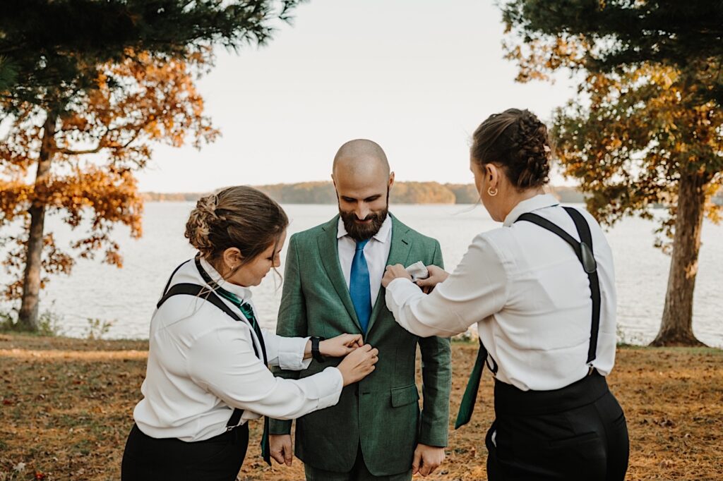 A groom stands and smiles while looking down as the two members of his wedding party adjust his suit pocket handkerchief and the button of his suit, behind them is a lake